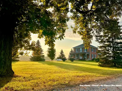 2023 2nd Place Photo - Magic hour glow on front yard with a tree in the foreground and brick farmhouse in the background, taken by Lori Hobin